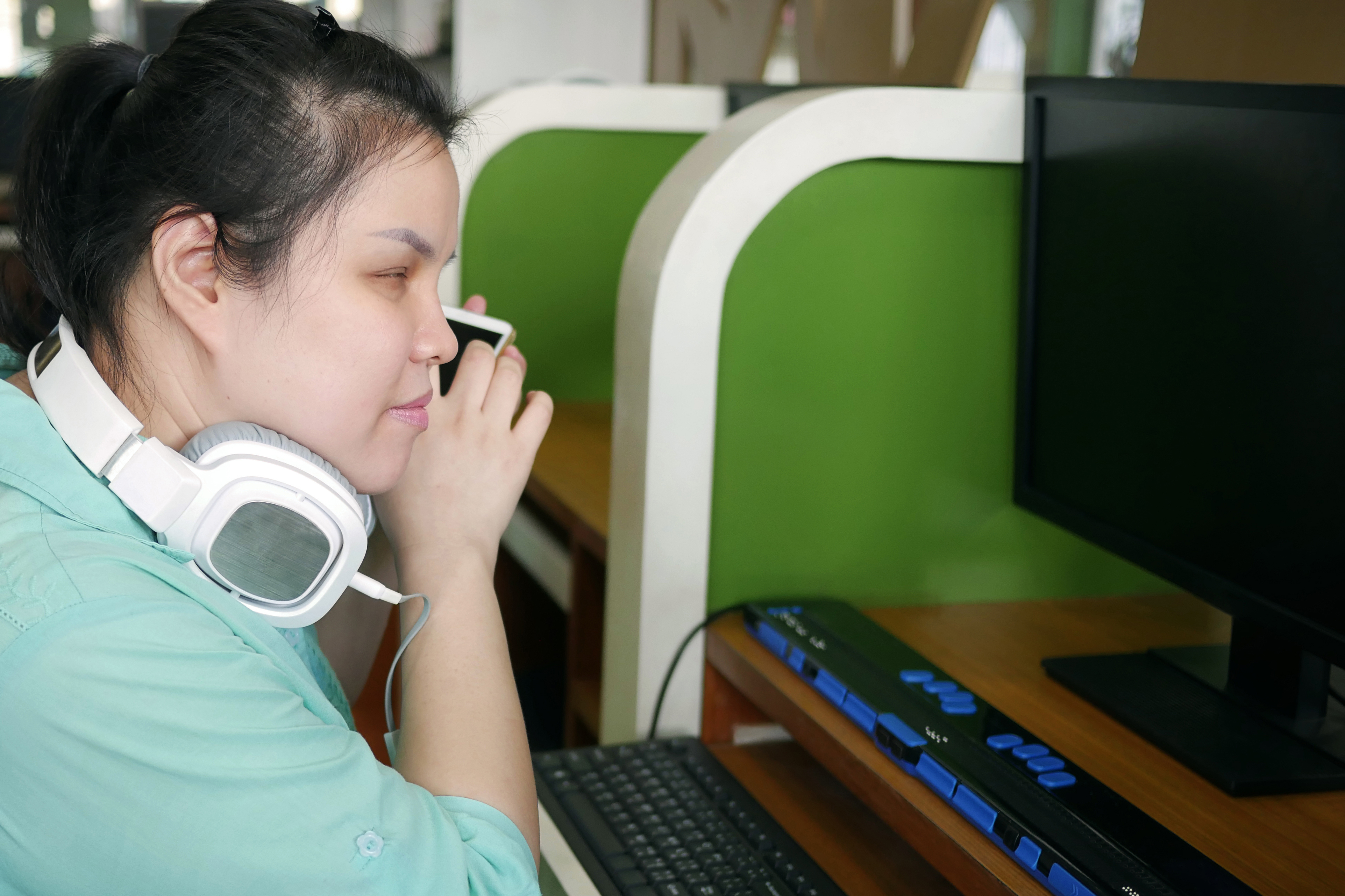 A young blind Asian woman viewed from profile, listening to her phone. She is wearing over the head headphones, and sitting in front of a computer with a refreshible display.