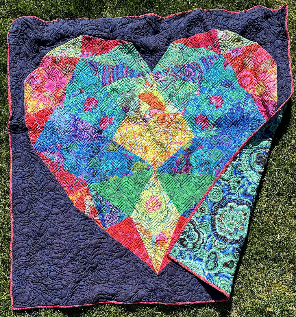 A 60 inch square quilt on the lawn. The quilt has a dark purple background and a patchwork heart shape occupies nearly the entirety of the quilt. The fabrics for the heart are a riot of Kaffe Fassett fabrics in reds, oranges, blues, yellows, and greens. The bottom right of the quilt is folded over showing a teal printed backing