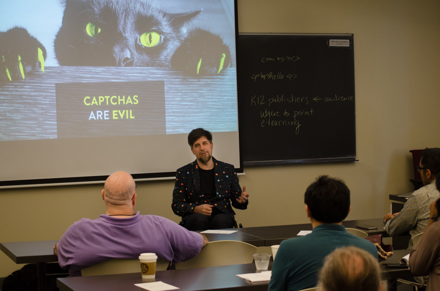 Nic Steenhout facing a crowd. A slide is shown behind Nic, with a cat reaching on top of a table and the words Captchas are Evil. Nic is using a wheelchair.