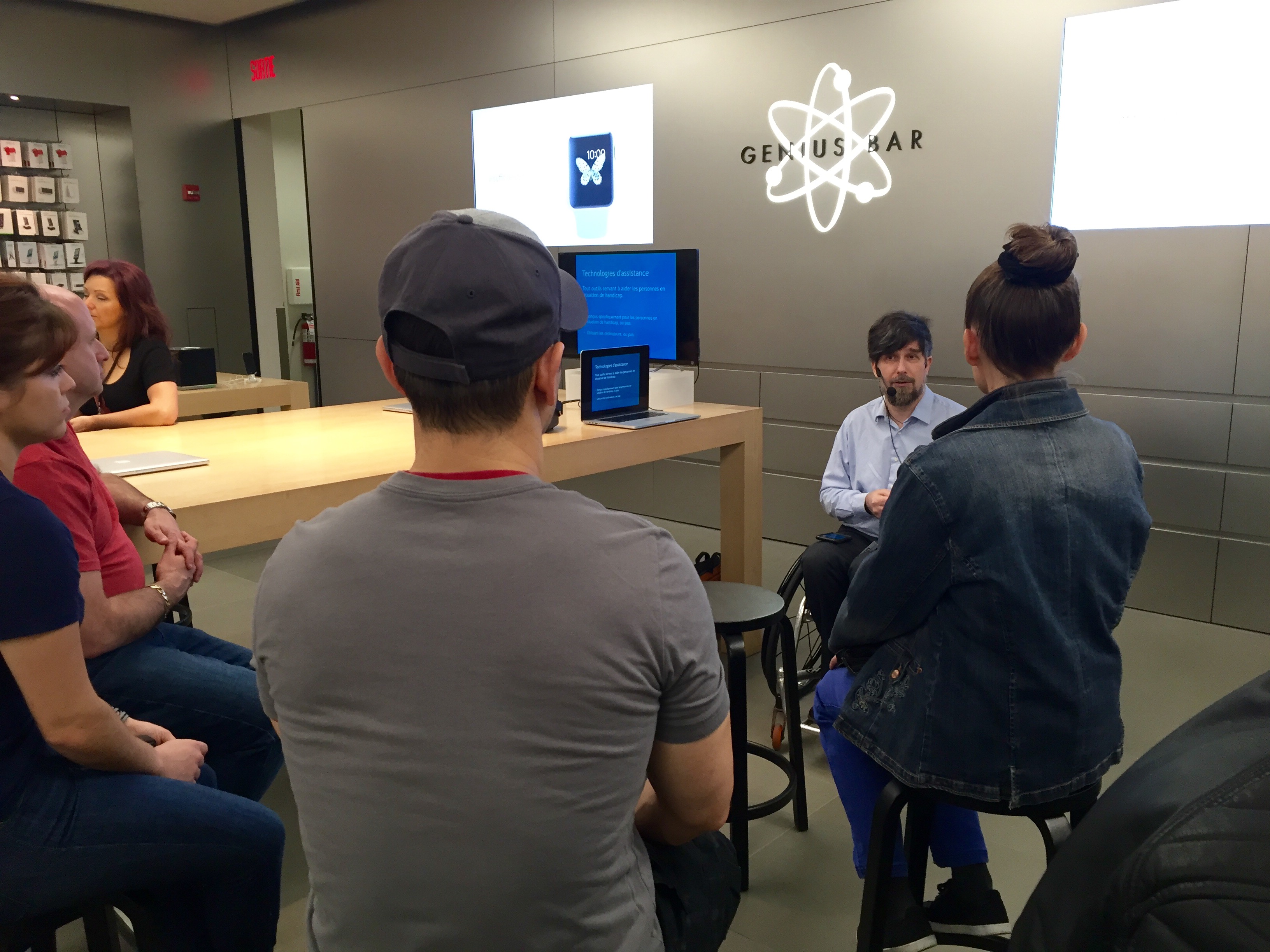 Nic Steenhout sitting in front of a group of people. The logo for Apple Genius Bar is on the wall behind Nic. Nic is using a wheelchair.