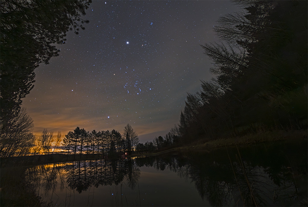 Night sky showing lots of stars. The mid ground of the image shows leafless trees in silhouette against a lighter colored sky. The foreground is a still lake with the trees and the stars reflected in it.