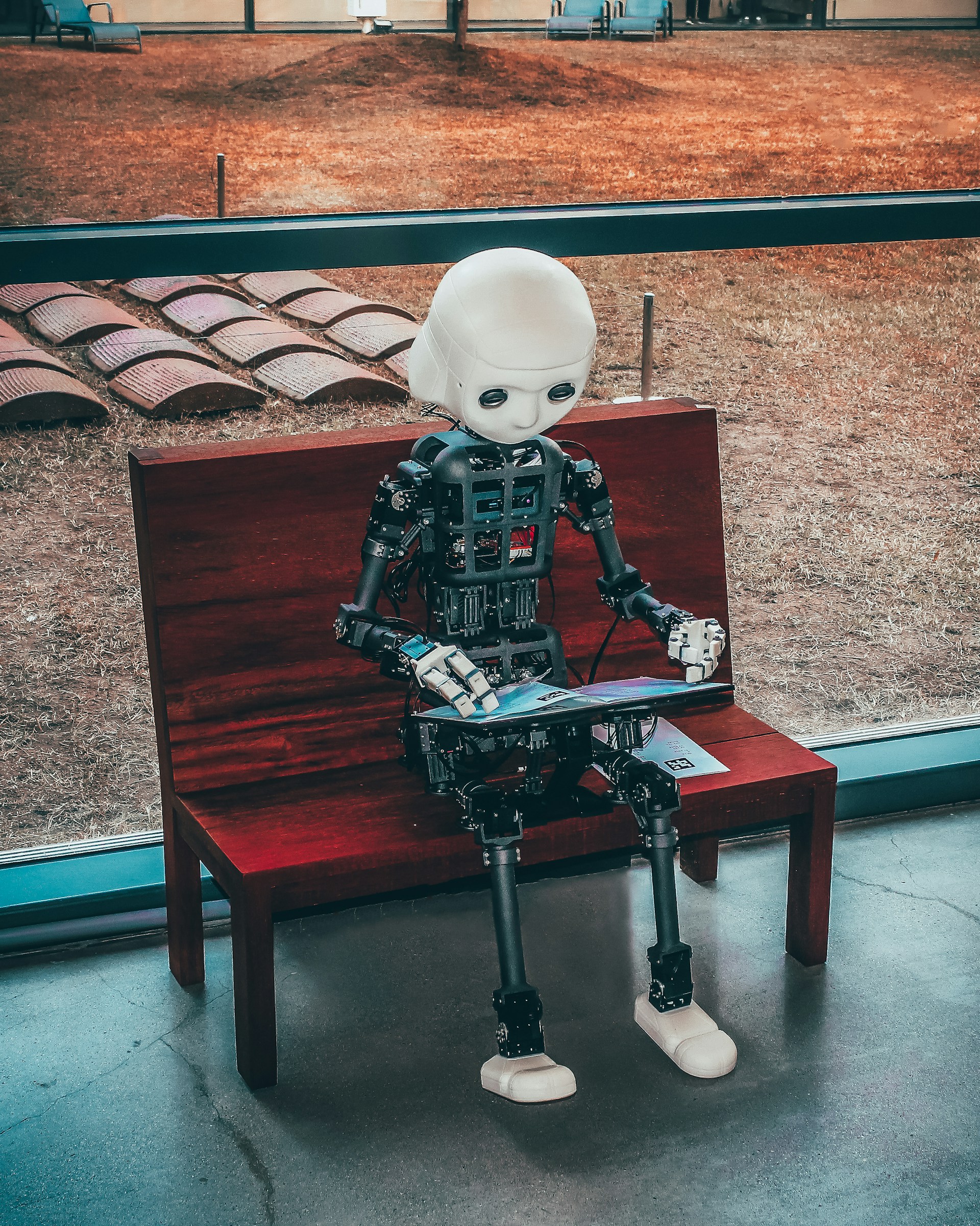 Human-shaped robot sitting on a wooden bench, reading a book.