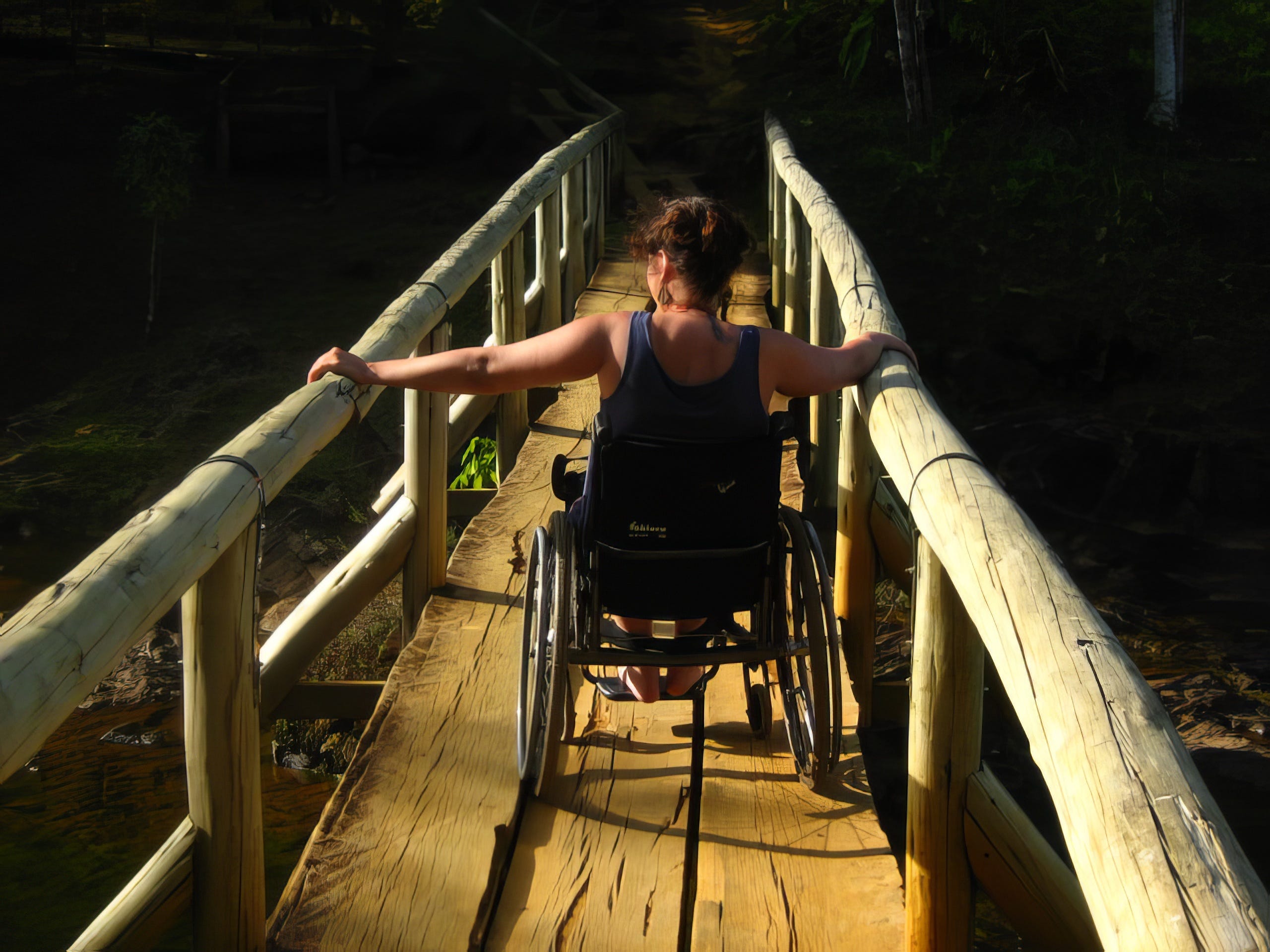 A wheelchair user viewed from the back, crossing a rickety small wooden bridge.